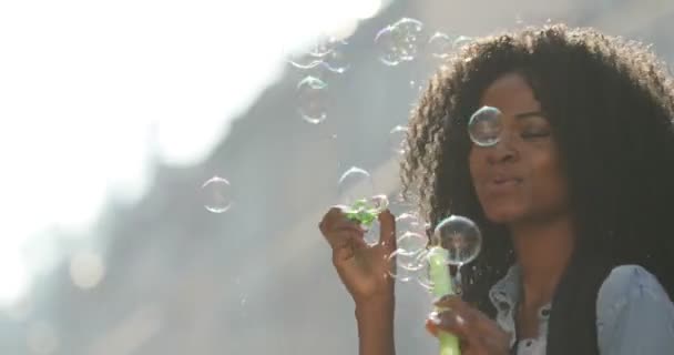Красива афро-американська дівчина з кучерявим волоссям дме мильні бульбашки і кружляє навколо під час прогулянки на сонячній вулиці. Портрет крупним планом . — стокове відео