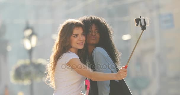 La vista de dos encantadoras amigas multiculturales enviando besos al aire mientras toman fotos usando el palo de selfie y balanceándose. Ubicación de la calle . — Vídeos de Stock