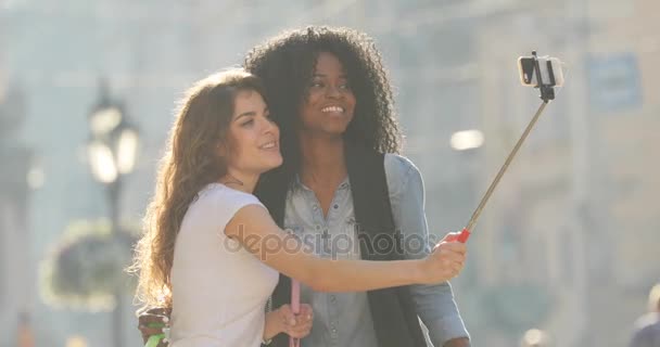 Deux copines multiculturelles ludiques sautent tout en prenant des photos d'elles-mêmes à l'aide du bâton de selfie. Emplacement de la rue . — Video