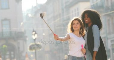 İki gülümseyen kız görünümünü Caddesi boyunca yürümek fotoğraflarını alıyor. Selfie sopa kullanıyorlar. Afro-Amerikan kız barış işareti gösteriyor.