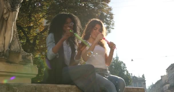 Dos encantadoras novias alegres están sentadas en la fuente y soplando las burbujas de jabón durante el atardecer. Uno de ellos es afroamericano. . — Vídeos de Stock