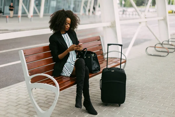 L'elegante vestito afro-a, viaggiatore erican è seduto sulla panchina e chatta tramite il telefono cellulare . — Foto Stock