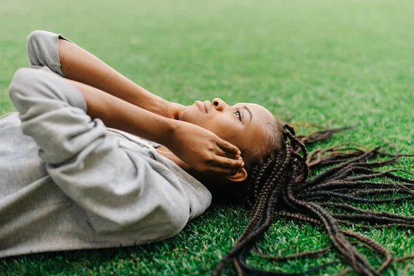 La bella sportgirl afro-americana sta ascoltando musica con le cuffie mentre è sdraiata sull'erba. Il ritratto ravvicinato . — Foto Stock