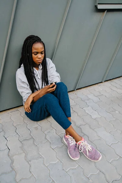 La vista anterior del triste adolescente afroamericano sentado en el suelo y apoyado en la pared. Ella está enviando mensajes a través del teléfono móvil . —  Fotos de Stock