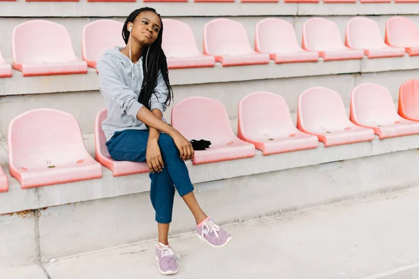 Adolescente afro-americano pacífica está ouvindo música em fones de ouvido enquanto se senta nos assentos cor de rosa no estádio . — Fotografia de Stock