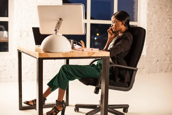 Mulher de negócios afro-americana sentada em sua cadeira na mesa e conversando ao telefone . — Fotografia de Stock