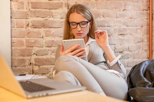 Žena sedí u stolu s notebookem na ní a držení tabletu v ruce. Žena nosí brýle a oblek. — Stock fotografie