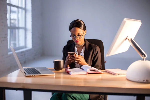 Uma jovem e atraente empresária afro-americana sentada no escritório com o telefone nas mãos. Laptop, PC e notebook colocados na mesa de madeira . — Fotografia de Stock