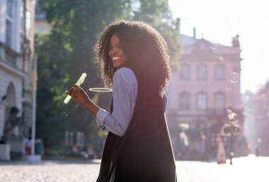 Kabarcık üfleyici tutan parlak bir gülümseme ile mutlu siyah genç kadın. Güneşli sokakta duran kadın.