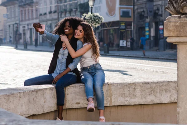 Selfie de duas meninas atraentes perto do monumento na bela cidade. Uma negra com a amiga. Meninas vestindo roupas casuais . — Fotografia de Stock