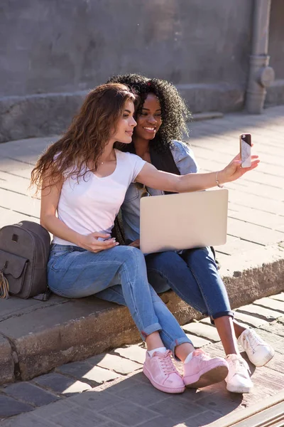Two happy young women sitting on the pavement and maling selfie. Black women holding laptop. Casual lifestyle concept.