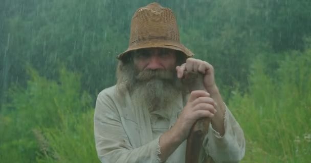 Un anciano agradable con barba gris larga y con ropa vieja está masticando mientras se apoya en su bastón bajo la lluvia. Retrato de primer plano . — Vídeo de stock