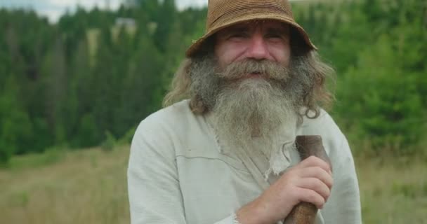 Nahaufnahme des lächelnden alten Mannes mit langen Haaren und Bart, der auf seinen Stock gestützt ist. Lage in den Bergen. — Stockvideo