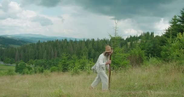 De wandeling van de oude grijze man in oude kleren houden het riet. Het prachtige uitzicht op de groene bergen. — Stockvideo