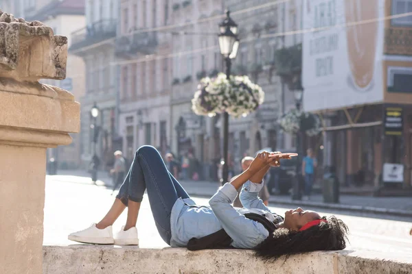 Mulher negra jovem atraente que se encontra perto do monumento no centro da cidade com o tablet em suas mãos. Mulher vestindo roupas casuais . — Fotografia de Stock