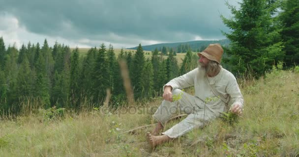 El senoir viajero en ropa vieja y con barba larga está sentado en la hierba y disfrutando de la vista de las montañas. Él toca y observa las flores . — Vídeo de stock