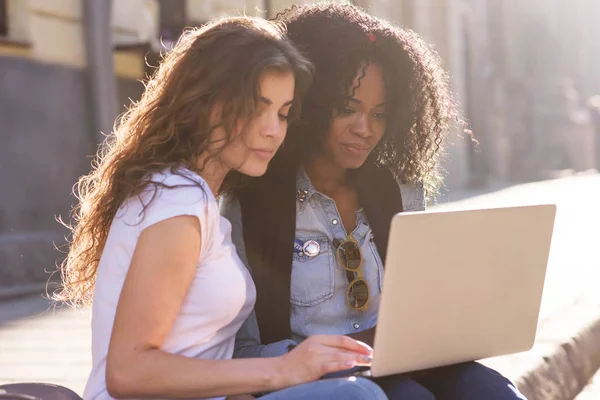 Close up tiro de duas meninas atraentes sentados na rua com o laptop e olhando para a tela do laptop . — Fotografia de Stock