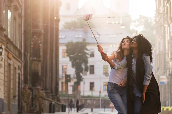 Duas meninas jovens e bonitas fazendo selfie em uma rua bonita. Uma rapariga é negra. Estilo de vida casual . — Fotografia de Stock