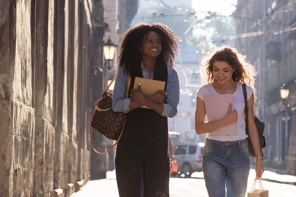 Mutlu öğrenciler ellerinde kitaplarla sokakta yürümek. Bir zenci kız beyaz arkadaşı. Güneşli bir gün ve güzel bir şehir. — Stok fotoğraf