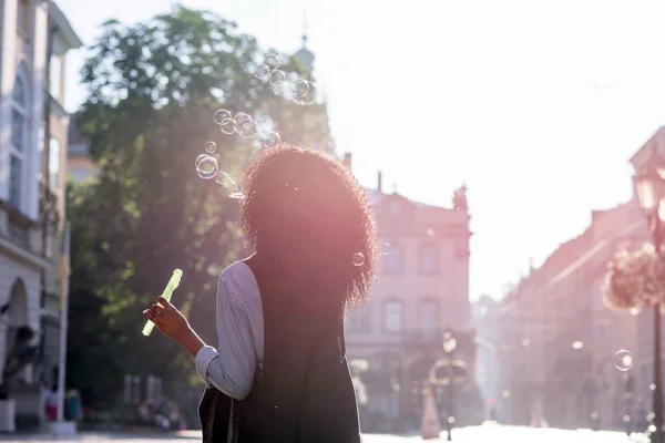 Femme noire aux cheveux bouclés debout dans la rue et soufflant des bulles. Concept de style de vie décontracté . — Photo