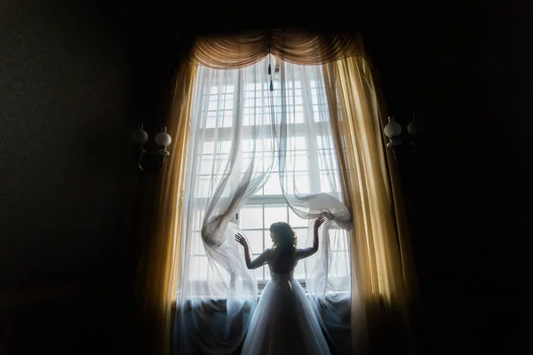 La mariée touche les rideaux tout en se tenant près de la fenêtre du palais. La vue arrière horizontale . — Photo