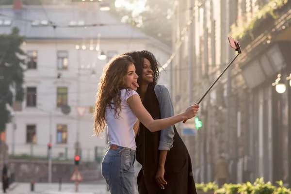 Zwei junge und attraktive Mädchen, die ein Selfie auf dem Vorplatz der schönen Stadt machen. ein Mädchen ist schwarz. — Stockfoto
