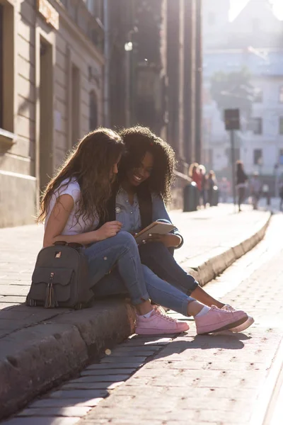 Dua gadis menarik duduk di trotoar dan melihat layar telepon. satu gadis african american. Indah kota di sekitar . — Stok Foto