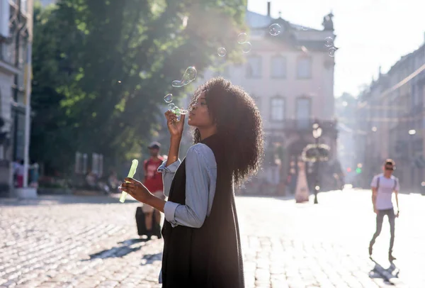 Joven y hermosa mujer afroamericana soplando burbujas en la calle. Buen tiempo y día soleado . — Foto de Stock