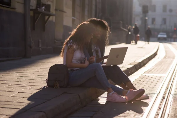 Attractive friends sitting on the pavement with the laptop and looking at the laptop screen. Twoo young girls. One girl is black. — Stock Photo, Image