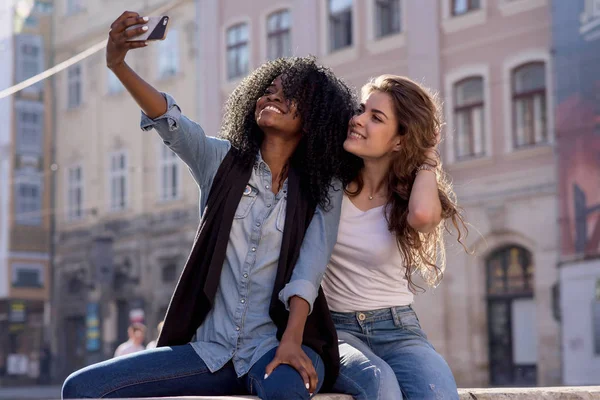 İki arkadaş selfie yapma gülümsedi. Kadınların rahat kıyafetler giyiyor. Siyah bir kadındır. Güzel mimari arka plan.