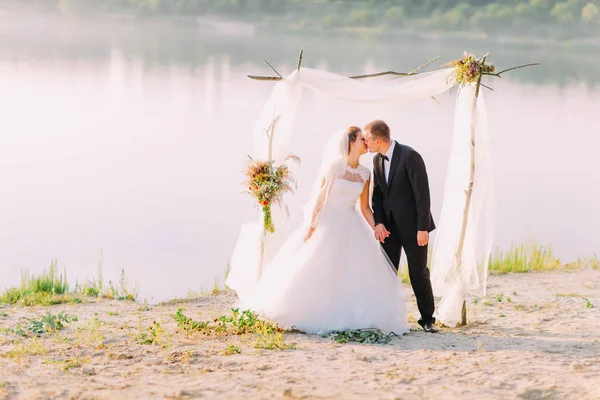 Horizontální pohled líbání newlywedsin přední oblouk s květinami umístěn v blízkosti řeky. — Stock fotografie