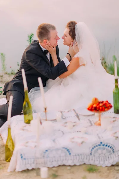 O retrato lateral sensível dos recém-casados acariciando uns aos outros rostos durante o piquenique de casamento perto do mar . — Fotografia de Stock