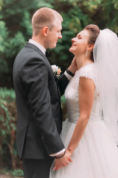 The side portrait of the smiling groom petting the face of the bride at the background of the park. — Stock Photo, Image