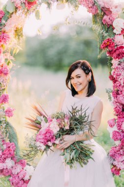 Büyük çiçek buketi ile mutlu gülümseyen gelin düğün peonies Arch güneşli parkta oturuyor.
