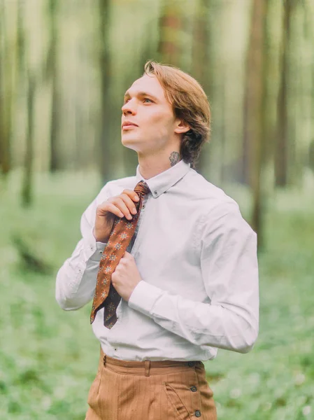O homem com cabelo loiro e tatuagem no pescoço com a camisa branca amarra sua gravata marrom e olha para o céu na floresta verde. Retrato lateral . — Fotografia de Stock
