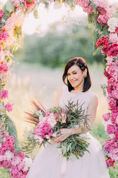 Büyük çiçek buketi ile mutlu gülümseyen gelin düğün peonies Arch güneşli parkta oturuyor.