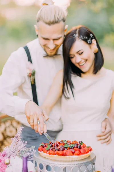 Das Nahaufnahme-Porträt des verschwommenen Brautpaares beim Anschneiden der Hochzeitstorte mit Kirschen und Erdbeeren. Das Hochzeitsessen findet im sonnigen Feld statt. — Stockfoto