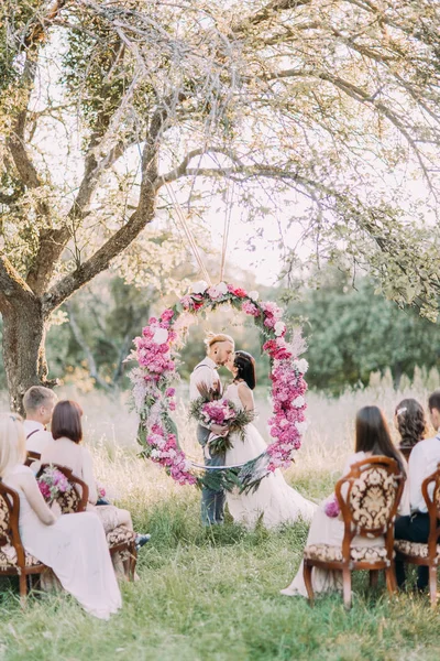 La foto verticale della bella cerimonia nuziale nella foresta soleggiata. Gli sposi baciare sono in piedi dietro l'arco peonie matrimonio mentre gli ospiti sono seduti sulle sedie . — Foto Stock