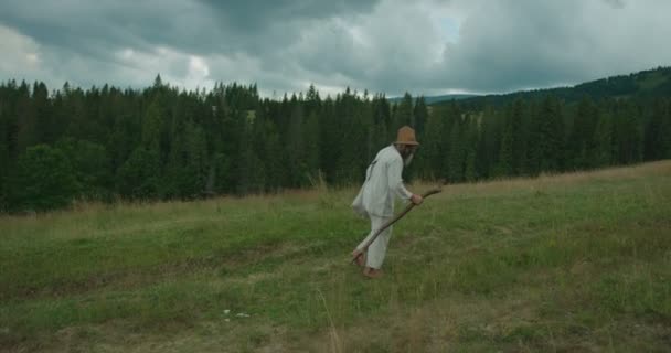 Plnohodnotné boční pohled starého muže v staré oblečení, s dlouhým vousem, chůze po zelené hory s pomocí hůl. — Stock video