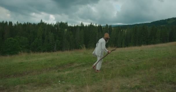 La vista laterale a tutta lunghezza del vecchio in abiti vecchi, con lunga barba che cammina lungo le verdi montagne con l'aiuto della canna . — Video Stock