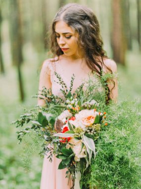 Yakın çekim portre renkli buket tutarak ve orman çim bakarak Pembe gelinlik uzun kıvırcık saçlı kadın.