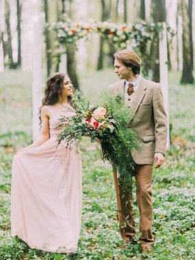 Güzel gülümsüyor yeni evliler yürüyüş ve arka plan beyaz kemer iki kapı kırmızı ve beyaz çiçeklerle süslenmiş şeklinde birbirlerine bakıyor. Damat buket tutuyor.