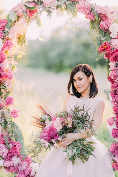 Το πορτρέτο της χαμογελαστός νύφη κρατώντας την ανθοδέσμη των λουλουδιών και να κάθεται στην αψίδα γάμου παιώνιες τοποθετούνται σε ηλιόλουστες πράσινο πάρκο. — Φωτογραφία Αρχείου