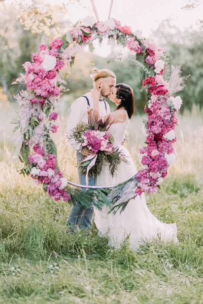O casal recém-casado está se beijando atrás do arco peônias de casamento na madeira ensolarada. Retrato de close-up . — Fotografia de Stock