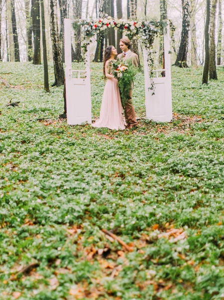 La foto vertical de los recién casados vestidos con ropa vintage y de pie bajo el arco en forma de puertas blancas decoradas con rosas rojas y blancas. El arco se coloca en el bosque verde . —  Fotos de Stock