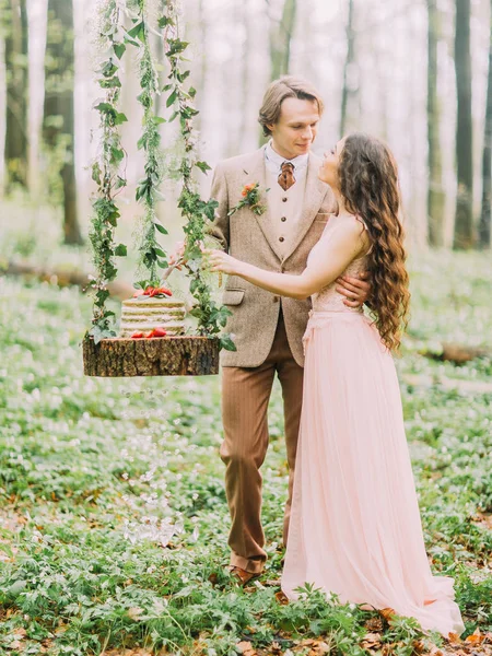 La photo verticale des jeunes mariés debout près de la souche suspendue avec le gâteau vert et blanc de mariage sur elle. Les heureux viennent de se marier dans la forêt printanière verte . — Photo