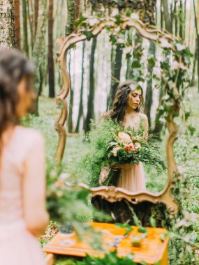 Dekore edilmiş görüntüde pembe elbiseli kadına çiçek ayna ile büyük renkli buket basılı tutup sağ tarafındaki ormanda bakarak.