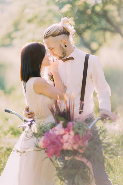 The close-up vertical photo of the lovely newlyweds carrying the bicycle with the bouquet of pink flowers at the background of the sunny forest. — Stock Photo, Image