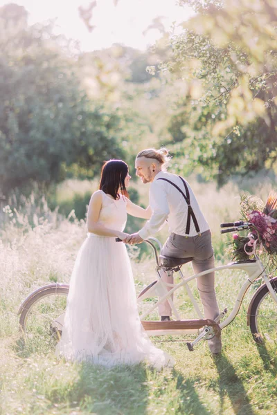 A foto vertical dos recém-casados na floresta ensolarada. A noiva no vestido branco está acariciando o ombro do noivo moderno sentado na bicicleta . — Fotografia de Stock