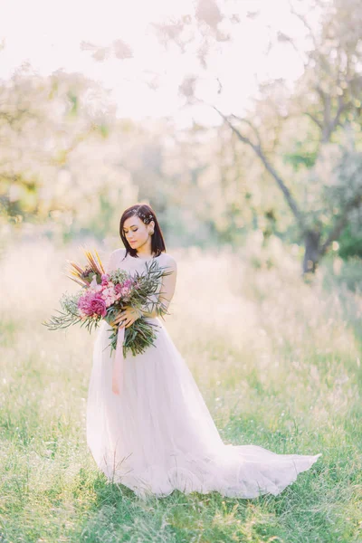 Η κάθετη φωτογραφία του την όμορφη νύφη στο Μακρύ γυναικείο φόρεμα, αξεσουάρ μαλλιών, κρατώντας το μεγάλο μπουκέτο με ροζ λουλούδια και περπάτημα στο πεδίο άνοιξη. — Φωτογραφία Αρχείου
